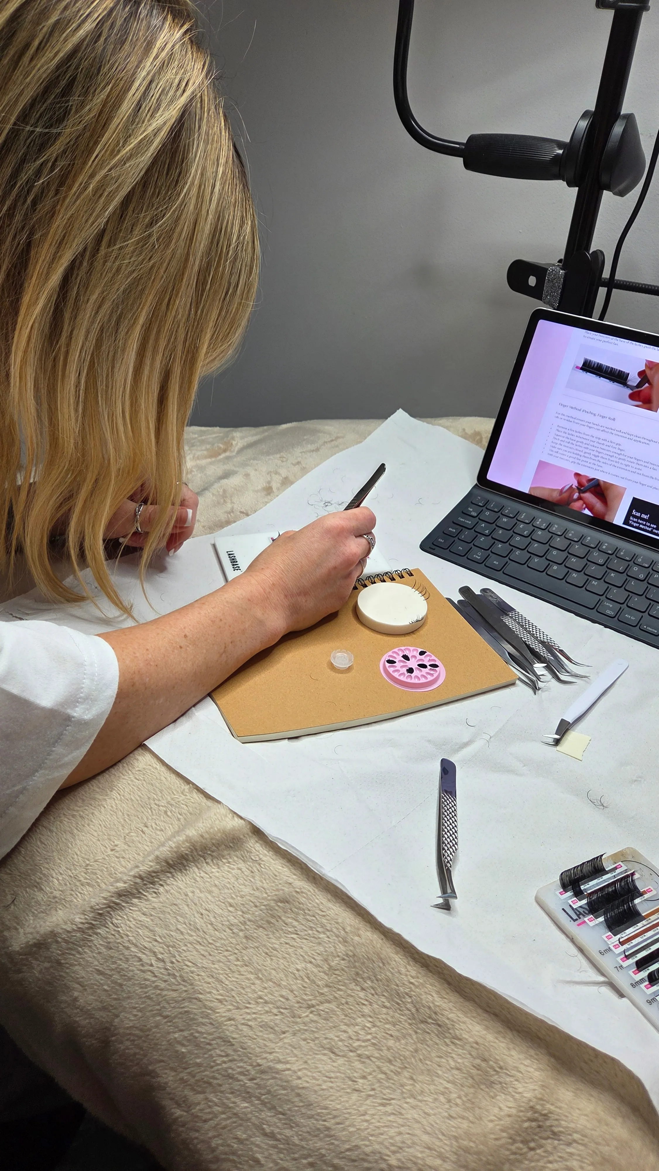 A student practising lash fans on a LashBase practice pad during a training day, manual and video guide open beside her